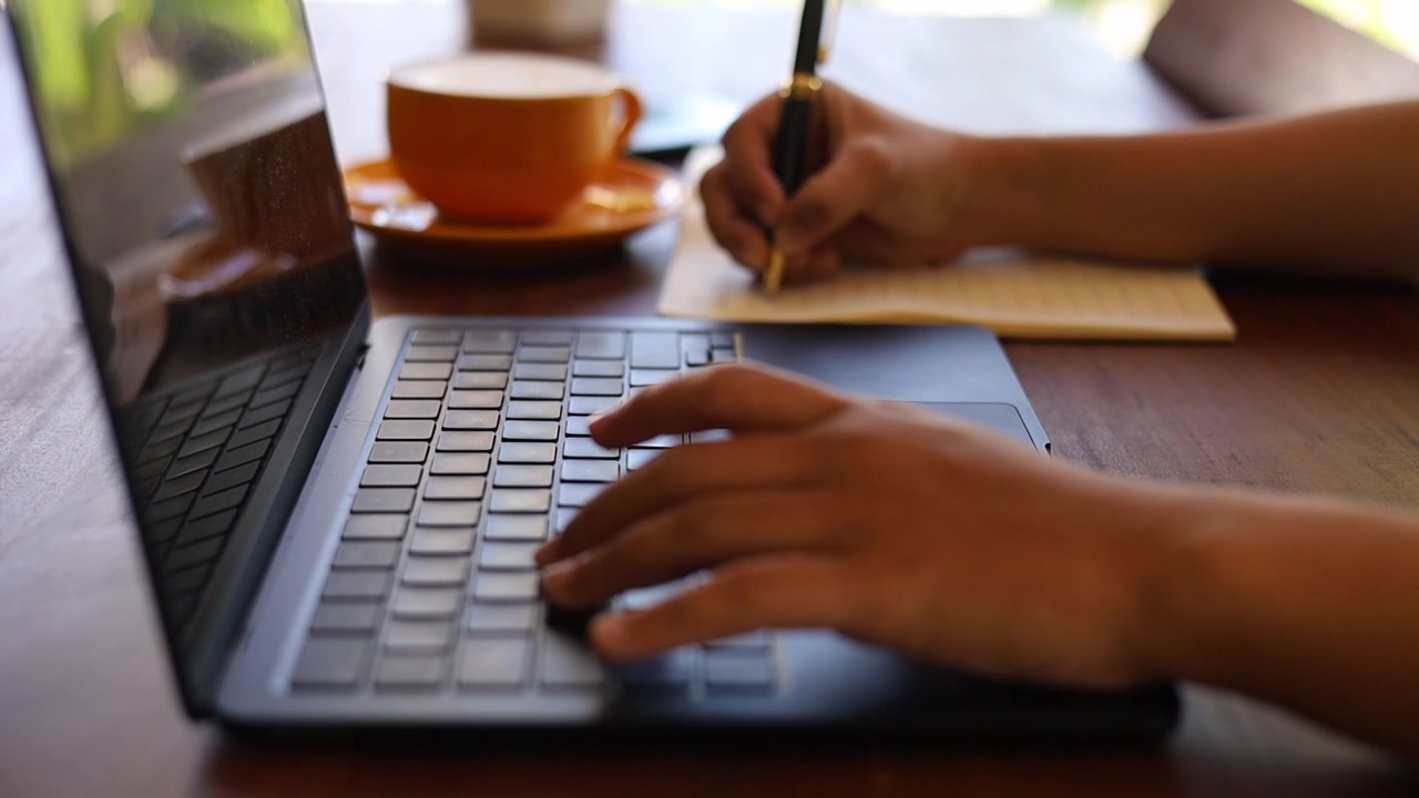 Primer plano de manos femeninas escribiendo en el teclado de un portátil, tomando notas, trabajando en una cafetería