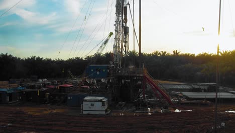 Cinematic-4K-Drone-Footage-of-Onshore-Drilling-Rig-equipment-structure-for-oil-exploration-and-exploitation-in-the-middle-of-jungle-surrounded-by-palm-oil-trees-during-sunset-and-high-oil-price