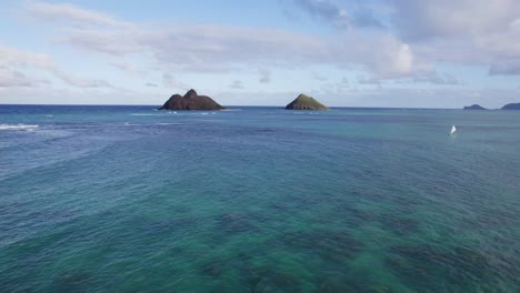 drone footage across the clear ocean water toward two small islands off the coast of oahu hawaii near lanikai beach with coral reefs showing through the blue green water