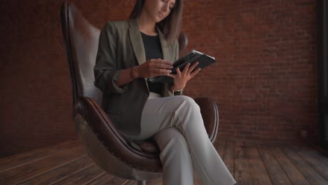 Brunette-sit-on-vintage-egg-chair-with-tablet-in-hands-near-red-brick-wall