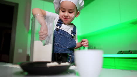 beautiful toddler kid in chef cap stands at the kitchen table. baby boy fills the black cup with flour. low angle view.