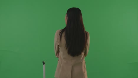 vista trasera mujer de negocios asiática viajera con equipaje cruzando los brazos y mirando a su alrededor mientras está de pie en el estudio de fondo de pantalla verde, durante la hora de la puesta del sol