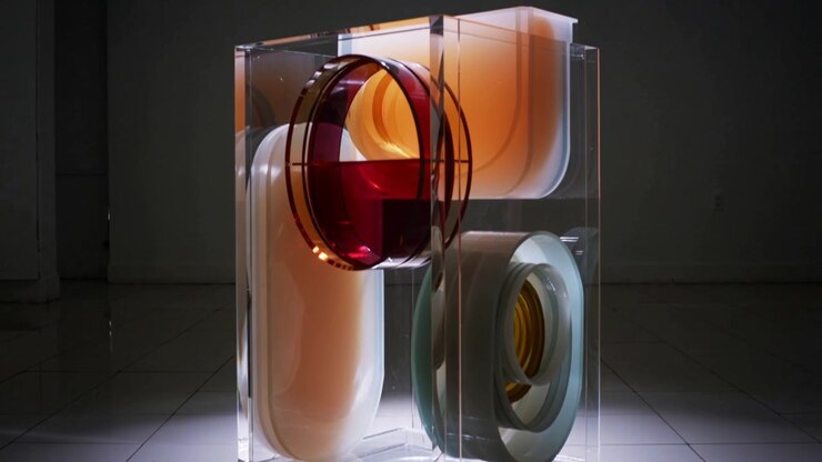 Telecamera che si allontana rivelando un cubo trasparente in una galleria in penombra, esponendo una sfera rossa, un pannello ambra e un anello grigio.