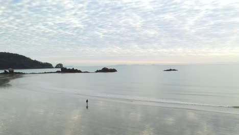 person walking alone on empty coastal bay beach at sunset, aerial arc shot