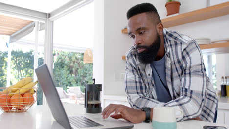 hombre afroamericano bebiendo café y usando una computadora portátil en una cocina soleada, en cámara lenta