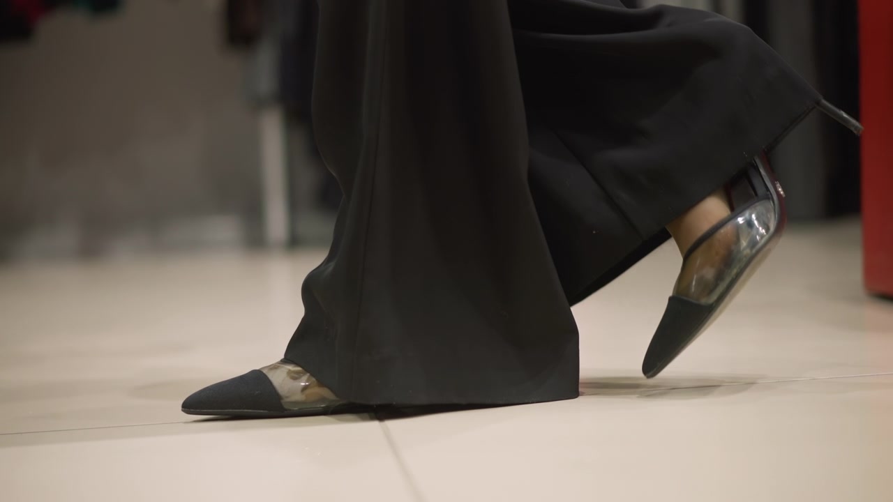 Primo piano dei piedi di una donna con scarpe nere a punta e tacchi trasparenti che cammina su un pavimento di colore chiaro. Il soggetto indossa pantaloni scuri a gamba larga e lo sfondo è un ambiente interno leggermente sfocato
