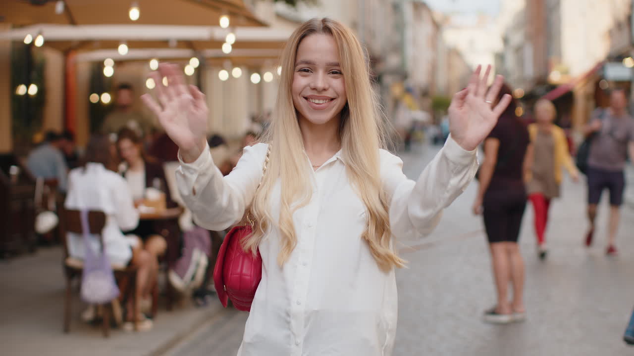 Young woman smiling friendly at camera waving hands hello hi greeting or goodbye in city street
