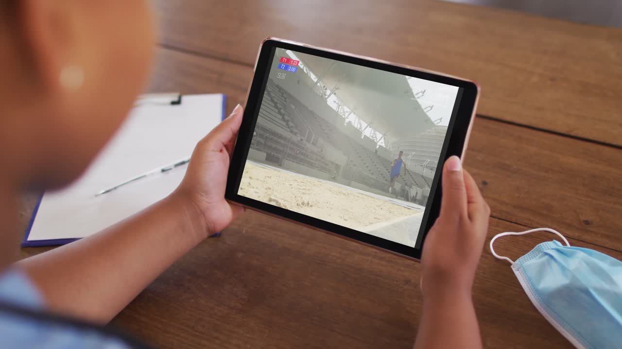 태블릿에서 육상 멀리 점프 이벤트를 시청하는 얼굴 마스크와 함께 테이블에 앉아있는 여성의 복합