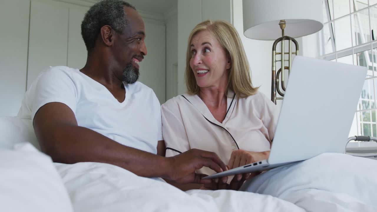 Diverse senior couple sitting in bed using laptop talking and laughing