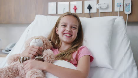 mother visiting daughter lying in bed in hospital ward and giving her teddy bear