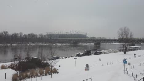 Drohnenvideo-Des-Warschauer-Nationalstadions,-Während-Ein-Zug-An-Einem-Verschneiten-Tag-über-Der-Weichsel-2-Auf-Einer-Brücke-Vorbeifährt