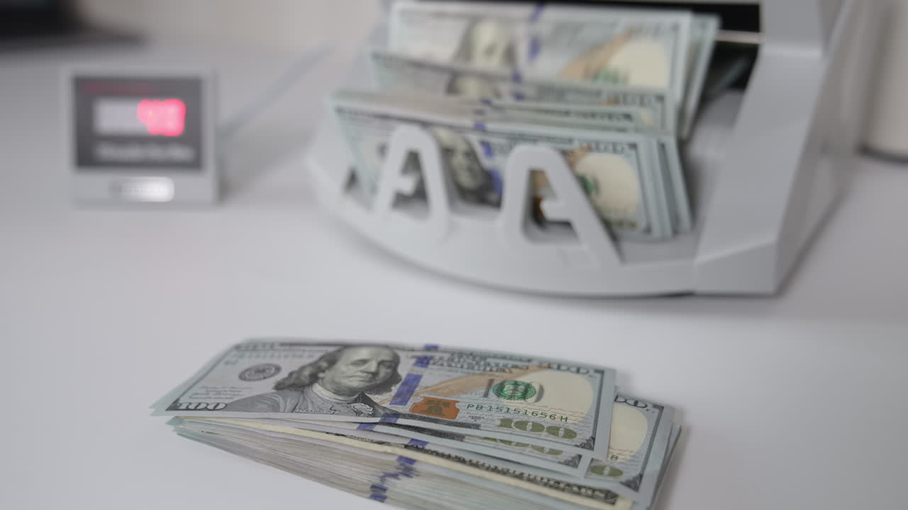 One hundred dollar banknotes on the table and in the cash counting machine. Male hand takes a wad of money showing them to the camera. Close up.