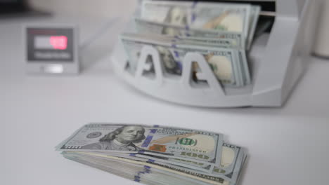 one hundred dollar banknotes on the table and in the cash counting machine. male hand takes a wad of money showing them to the camera. close up.