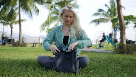 hermosa mujer delgada con el cabello largo rubio en camisa verde se sienta en el suelo y el uso de teléfono inteligente sobre el fondo del parque. niña en la pantalla cuadrada tocando y sonriendo