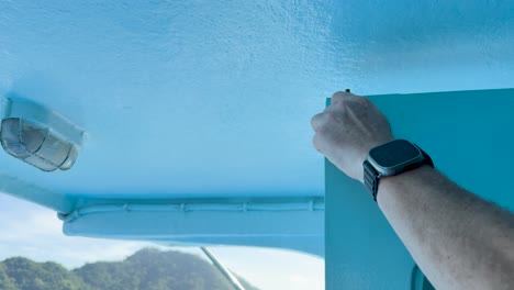 A person secures a blue metal door on a boat in Phuket, Thailand. Bright daylight illuminates the scene with scenic ocean views