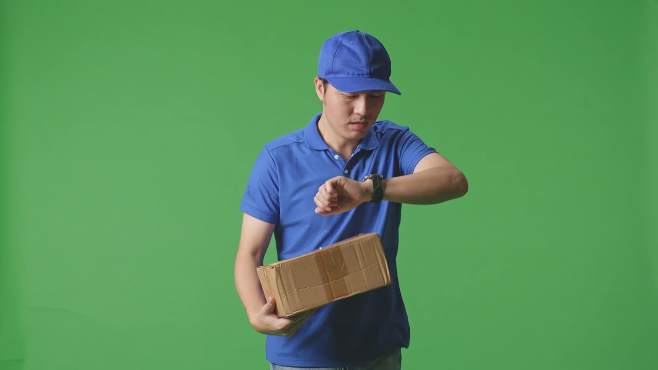 Asian Male Courier In Blue Uniform Checking Watch And Running While Delivering A Carton On Green Screen Background In The Studio