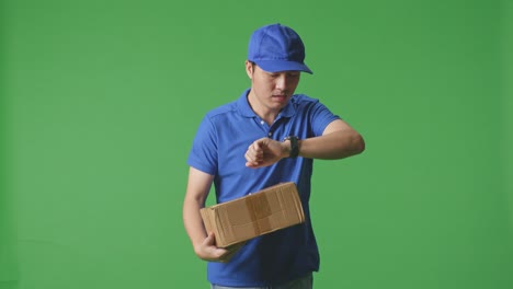 mensajero masculino asiático en uniforme azul comprobando el reloj y corriendo mientras entrega una caja de cartón en el fondo de la pantalla verde en el estudio
