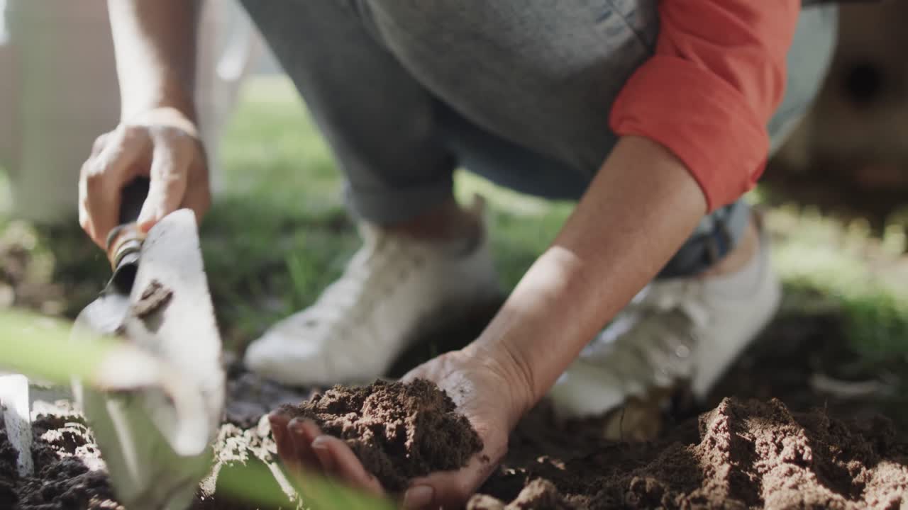 Dettaglio della parte inferiore di una donna anziana caucasica che prepara il terreno con le mani e una cazzuola in giardino, al rallentatore.