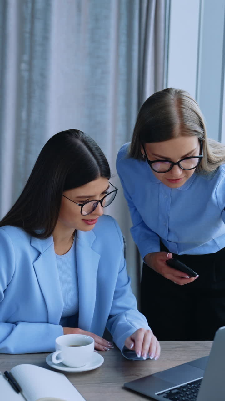 Business communication in the modern office. Female colleagues looking at laptop communicating and smiling. Blurred city panorama in window at backdrop. Vertical video