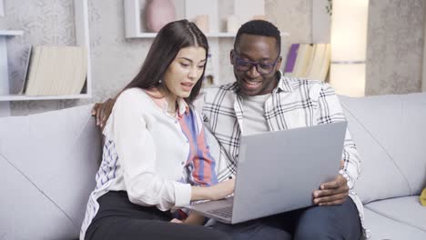 una feliz pareja casada multiétnica comprando en línea desde una computadora portátil en casa y son felices.