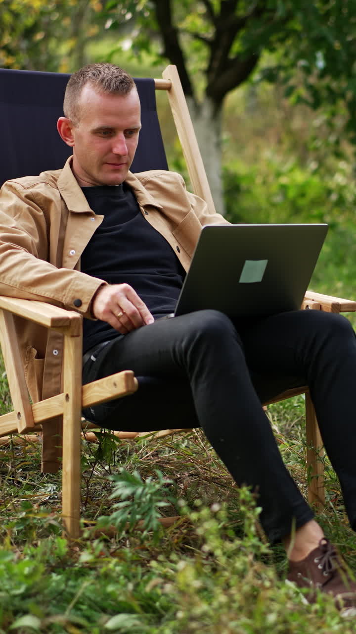 Calm relaxed freelancing man sitting in apple garden with laptop on his knees. Caucasian male working remotely online from nature. Vertical video