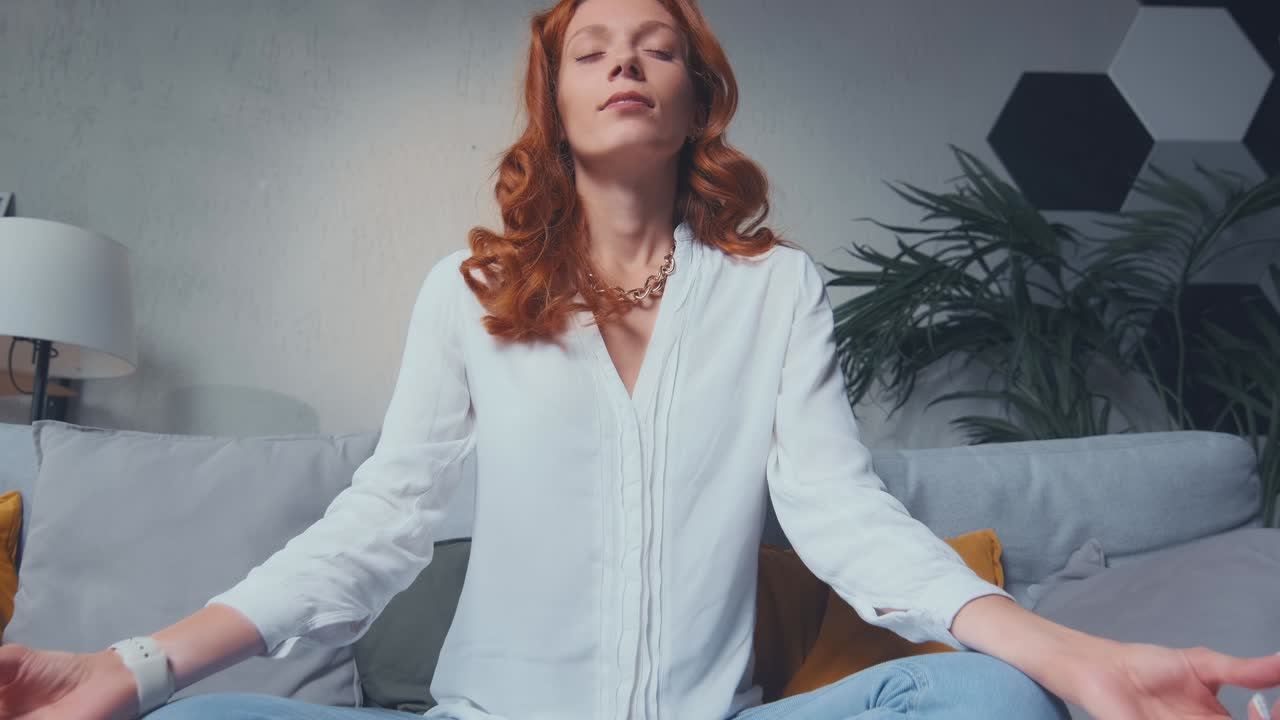 Red haired woman making mudra gesture sitting in lotus position on couch at home