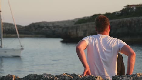 slow motion of man walking and sitting on a cliff watching a sailing yacht