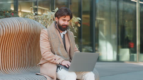 overjoyed happy young man working on laptop netbook screaming in delight on bench in downtown city
