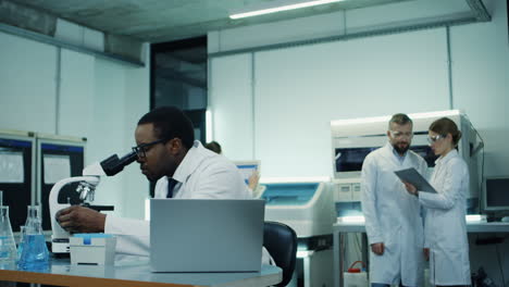 Joven-Científico-Con-Túnica-Blanca-Y-Anteojos-Trabajando-En-La-Computadora-Portátil-Y-El-Microscopio-Sobre-La-Investigación-Mientras-Su-Compañera-De-Trabajo-Viene-Con-Una-Tableta-Y-Discuten-Algunos-Resultados