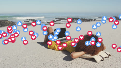 animation of red heart love and like digital icons over woman using laptop on beach