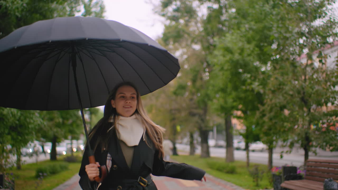 Pretty woman running happily under umbrella on windy autumn day, hair blowing in breeze, dressed in black trench coat and white hoodie, moving along city sidewalk with blurred cars and trees in background