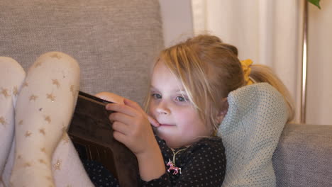 young girl lying on the couch playing with tablet device as screen reflects upon her face