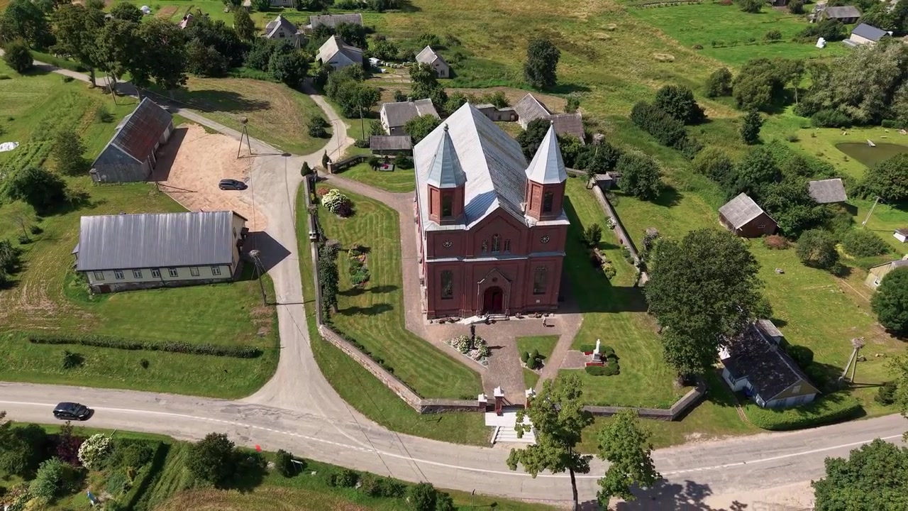 Drönarvideo visar en slående röd tegelkyrka med två spiror i en lummig lantlig by i Vilnius, Litauen. Solbelysta fält, pittoreska hem och panoramautsikt framhäver fridfull sommarskönhet.