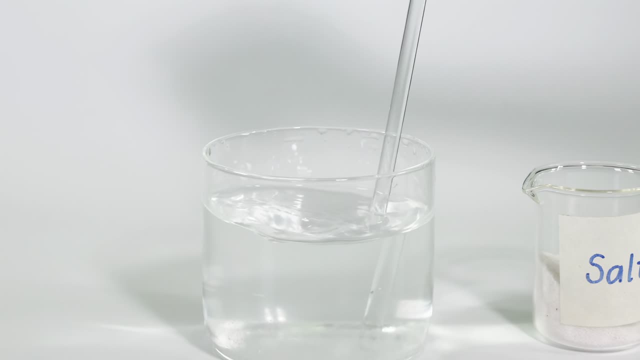 A glass rod stirs clear water in a beaker next to a labeled salt container.