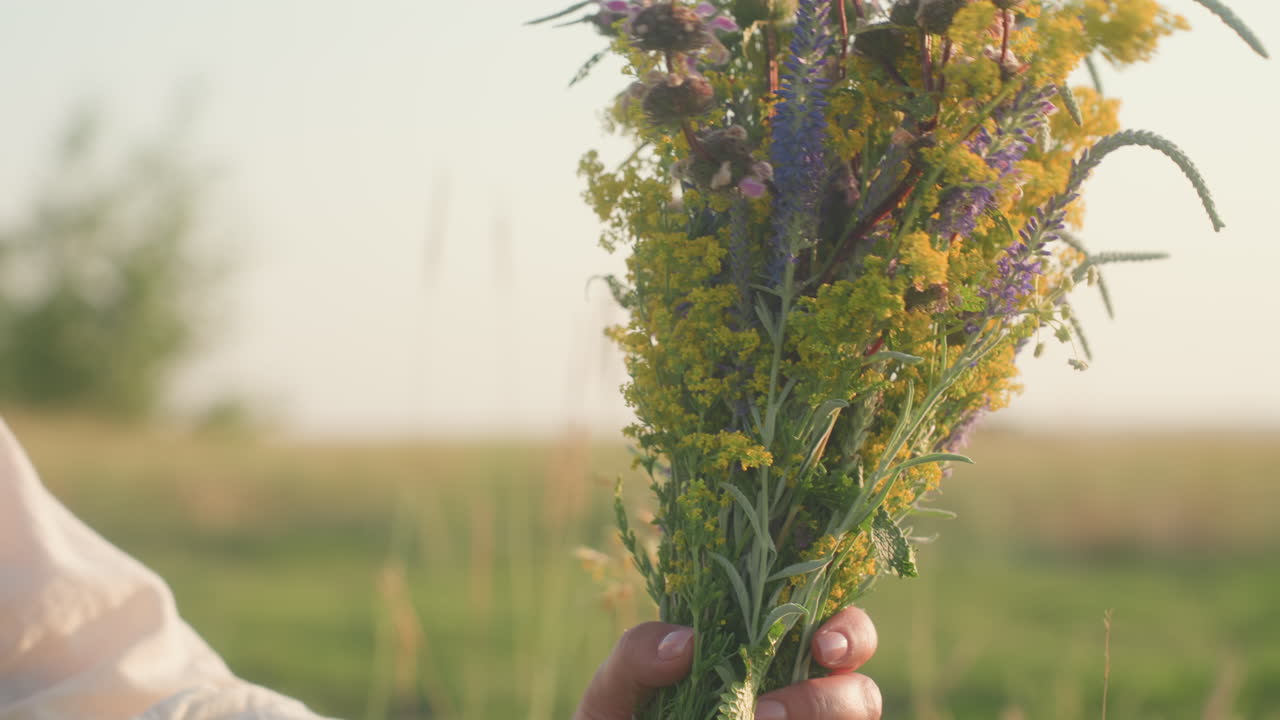 Primo piano di una donna con camicia bianca e vestito verde che tiene in entrambe le mani fiori di campo gialli e misti sotto la calda luce del sole, mentre sistema un bouquet in un tranquillo campo verde con uno sfondo sfocato e morbido.
