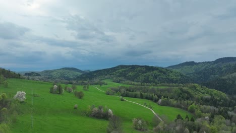 Descubra-La-Belleza-De-La-Primavera:-Un-Dron-Se-Desliza-A-Baja-Altura-Sobre-Huertos-Florecientes,-Prados-Y-Un-Granero-Abandonado,-Todo-Ello-Bajo-Espectaculares-Nubes,-Mientras-Una-Carretera-Sin-Pavimentar-Serpentea-A-Través-Del-Paisaje.