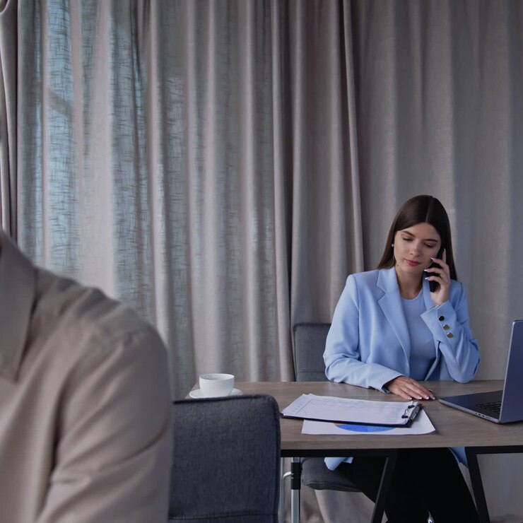 Young lady with dark long hair takes phone and starts conversation. Colleagues at the next desk speaking and using gadgets