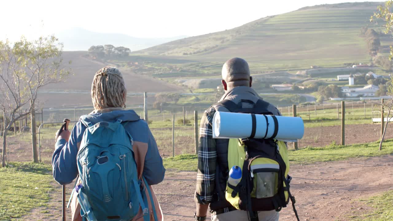pareja de ancianos afroamericanos con mochilas caminando en la naturaleza soleada, cámara lenta