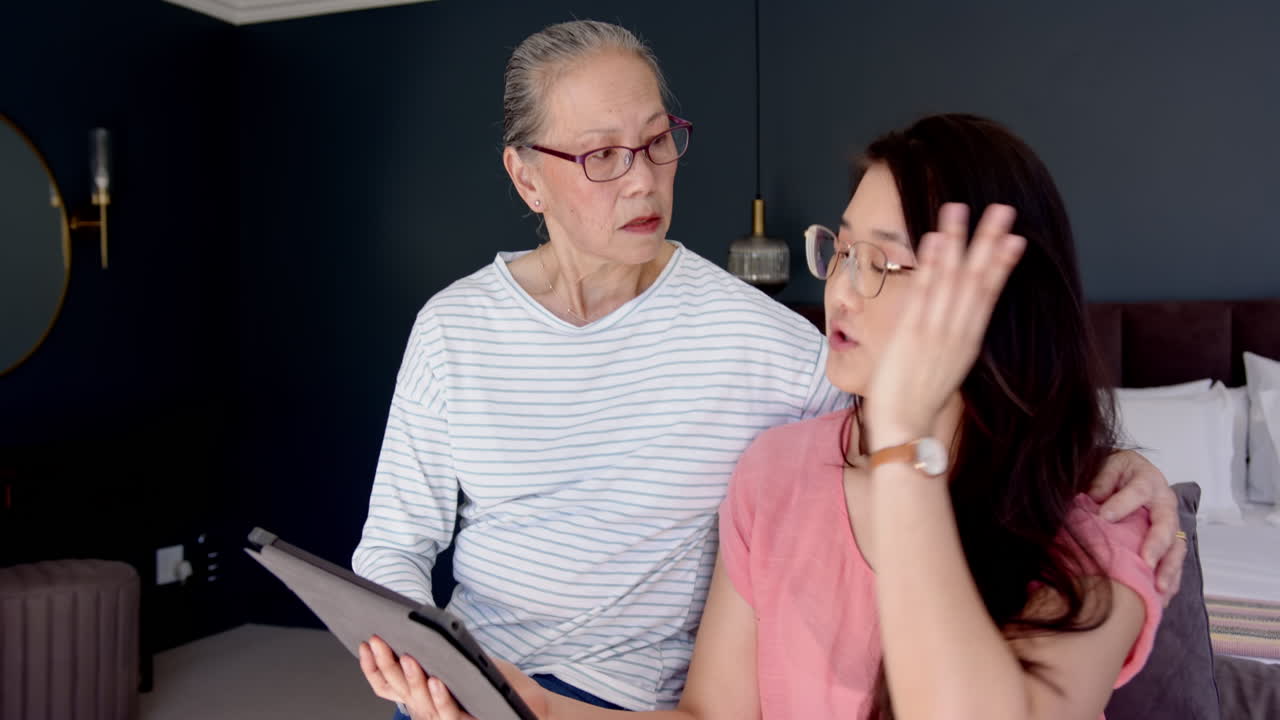Using tablet, Asian grandmother and granddaughter discussing important topic in living room