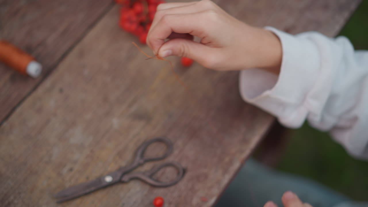 Closeup white hands threading red berries with thread and scissors on wooden table, delicate DIY garland making, cottage craft vibe, careful focused motion, tactile slow craft process, meditative