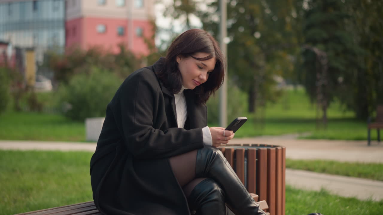 dama profesional en abrigo negro y cuello alto blanco, centrada en el teléfono con una sonrisa sutil, piernas cruzadas, sentada en un parque con un edificio residencial en el fondo