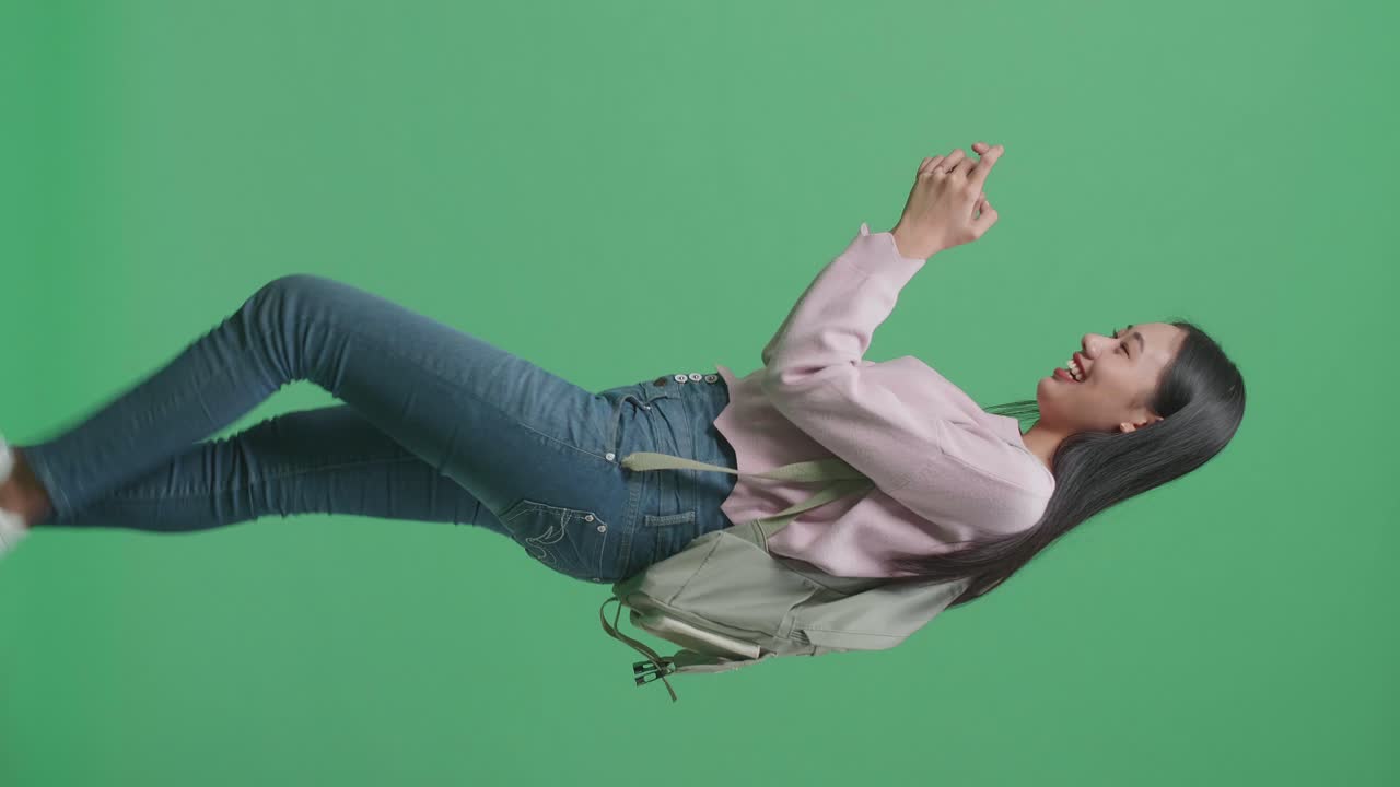 vista lateral de una estudiante asiática emocionada jugando a un juego móvil mientras camina en el estudio de pantalla verde