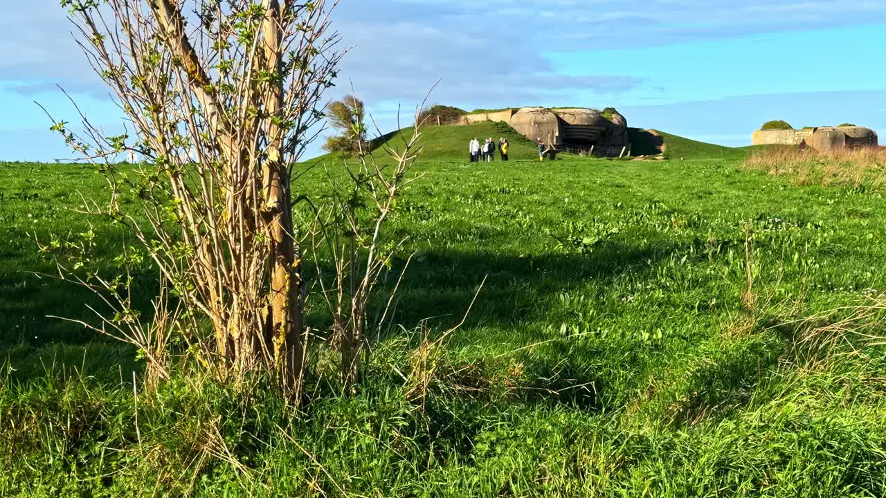 Laajakuva historiallisesta saksalaisesta tykistöpatterin kasematista Longues-sur-Merissä turistien kanssa vihreän rannikkokentän poikki sinisen taivaan alla Normandiassa Ranskassa