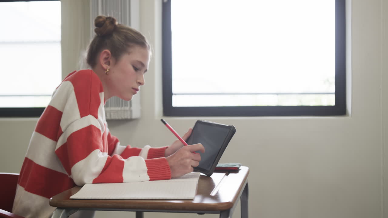 Young girl focusing on tablet and writing notes in classroom during study, at school, copy space