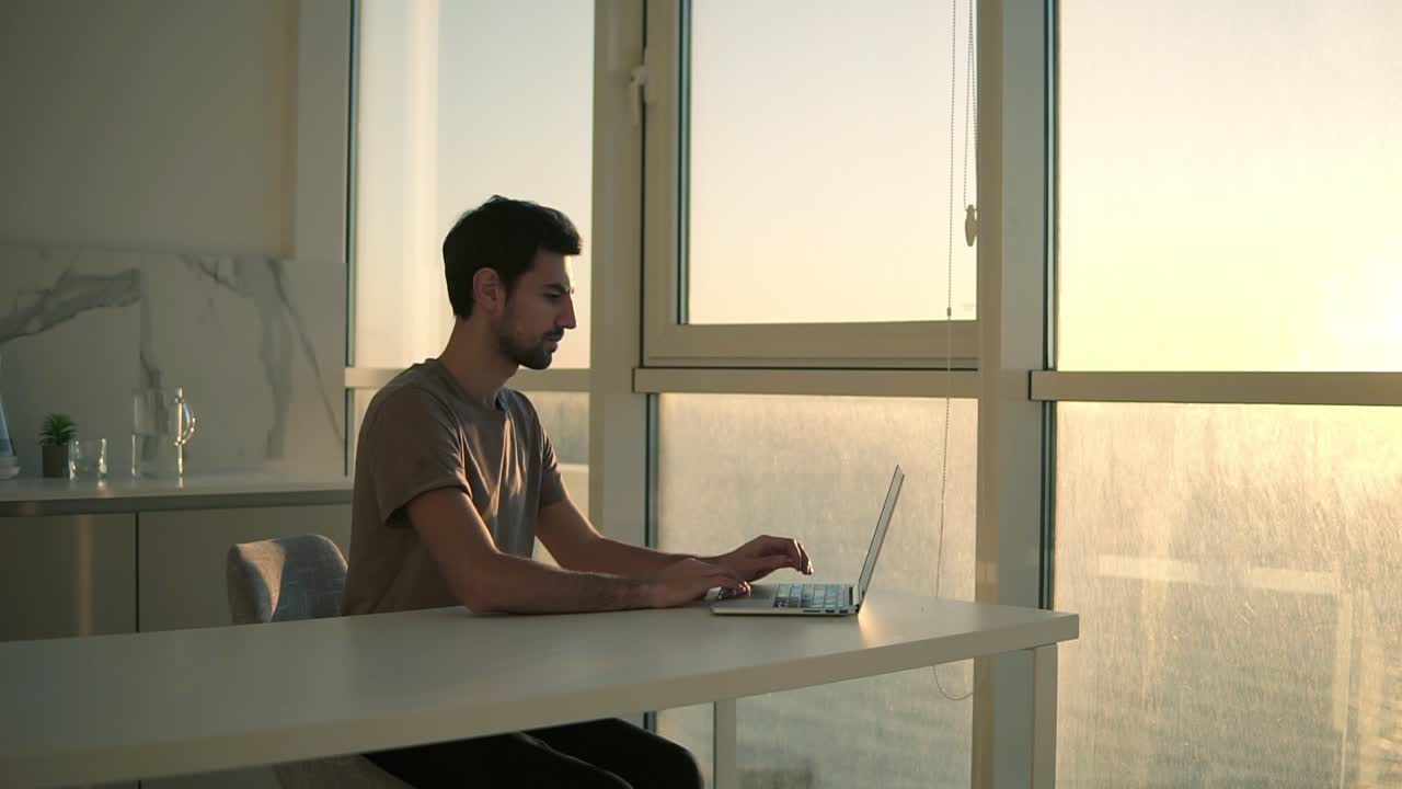 European dark haired man, freelancer opens laptop for work. Young guy sitting close to panoramic window on studio kitchen - focused on working with laptop and making difficult task. Slow motion