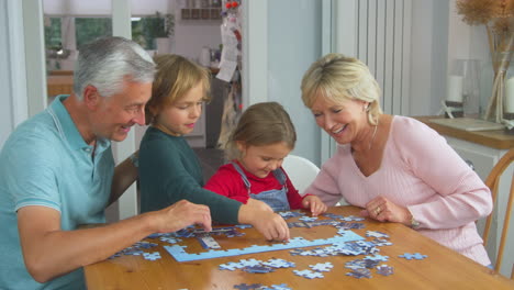 grandchildren with grandparents sit around table at home doing jigsaw puzzle together