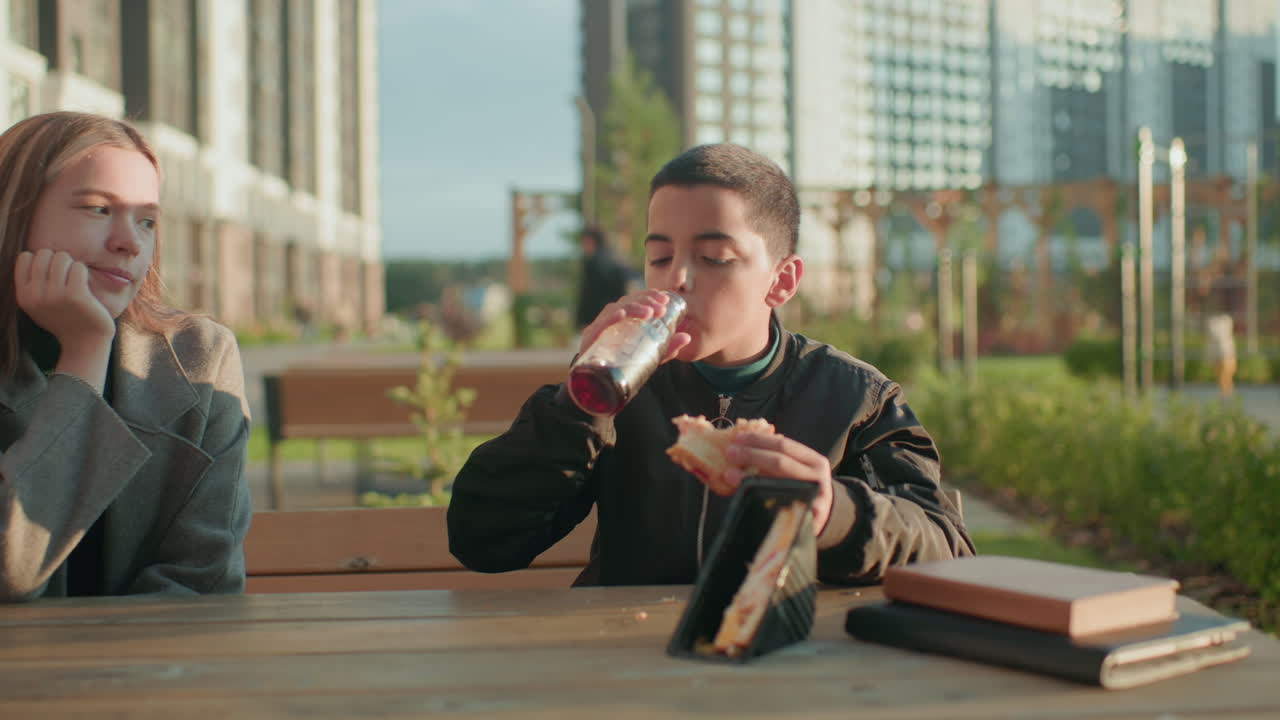 Guardian watching kid eating shawarma at outdoor wooden table with juice bottle, sandwich pack, and books, while someone passes in background