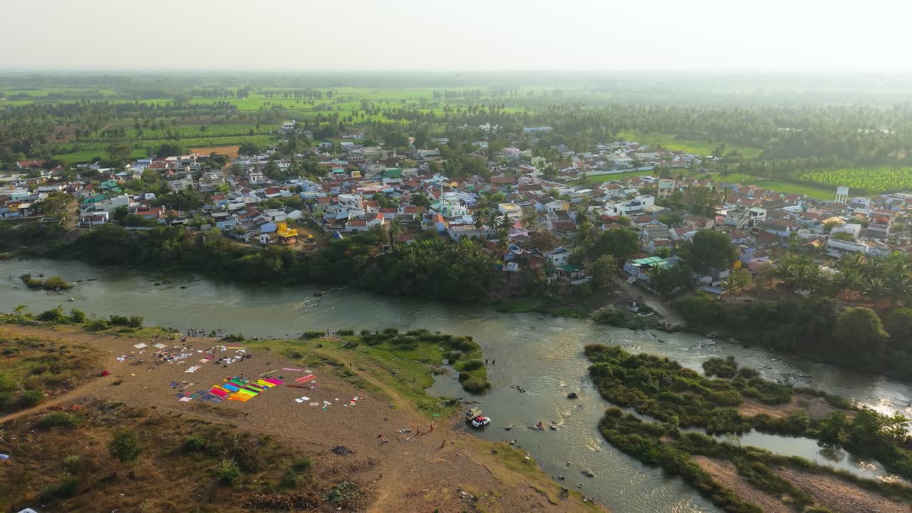 Vista aerea cinematografica di un villaggio rurale lungo il fiume, persone che lavano i vestiti e puliscono i veicoli lungo la riva, che mostra la vita rurale quotidiana, il flusso calmo dell'acqua e la tranquilla campagna circostante
