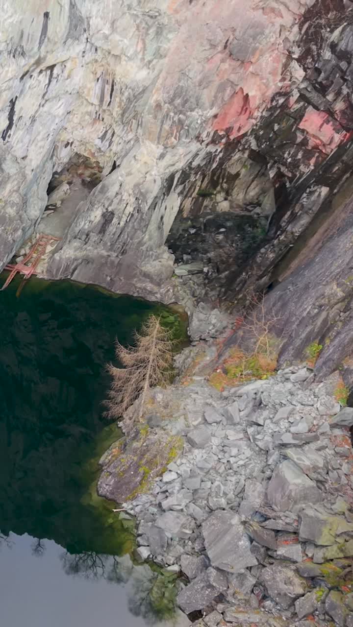 Discesa verticale di un drone sopra la cava di Hodge Close, con vista sul lago e sulle pareti di ardesia. Durante la discesa del drone, diventano visibili piccole grotte nella parete, rivelando la profondità impressionante e i dettagli robusti della cava.