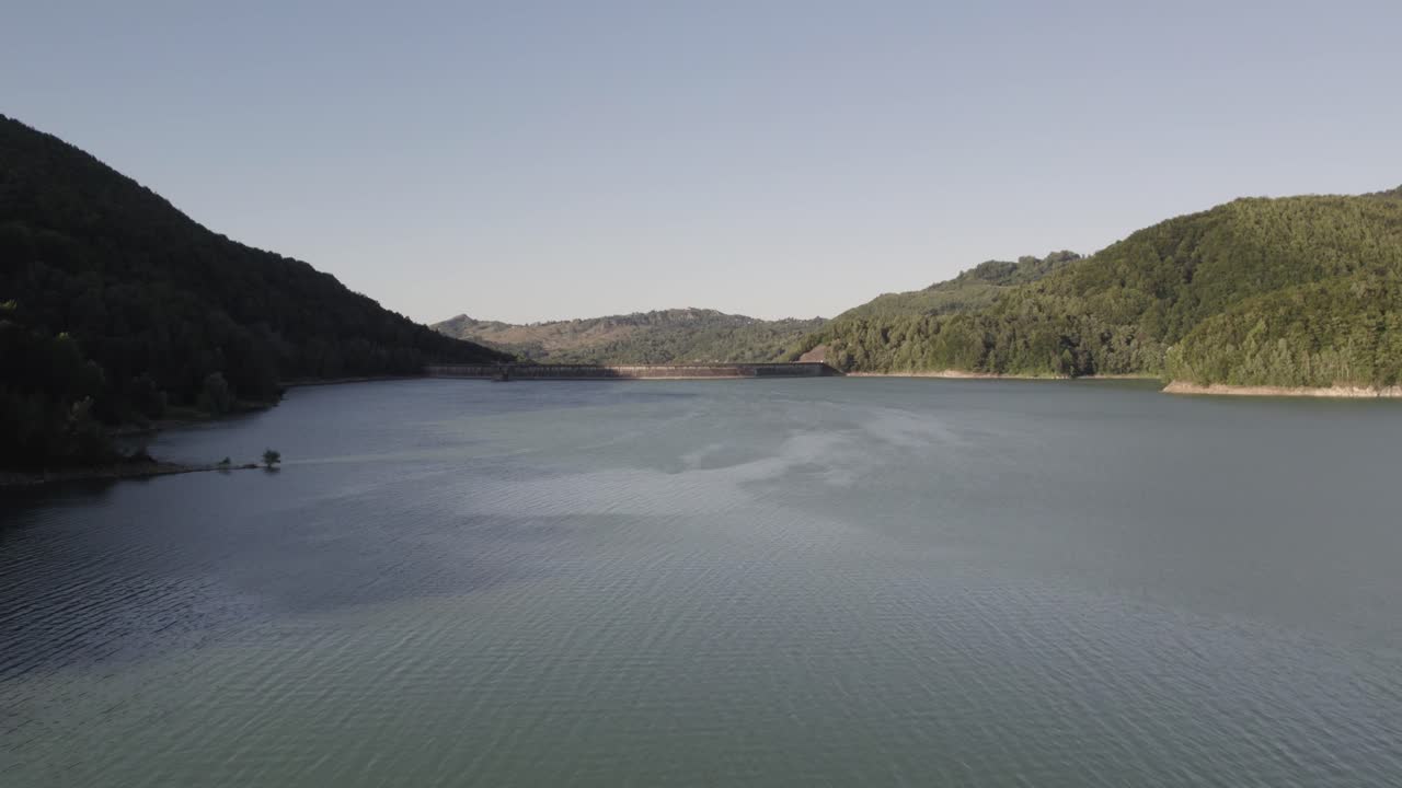 toma aérea, drone acercándose lentamente a una presa al final de un lago en rumania, volando sobre un enorme lago entre verdes colinas cubiertas por árboles forestales